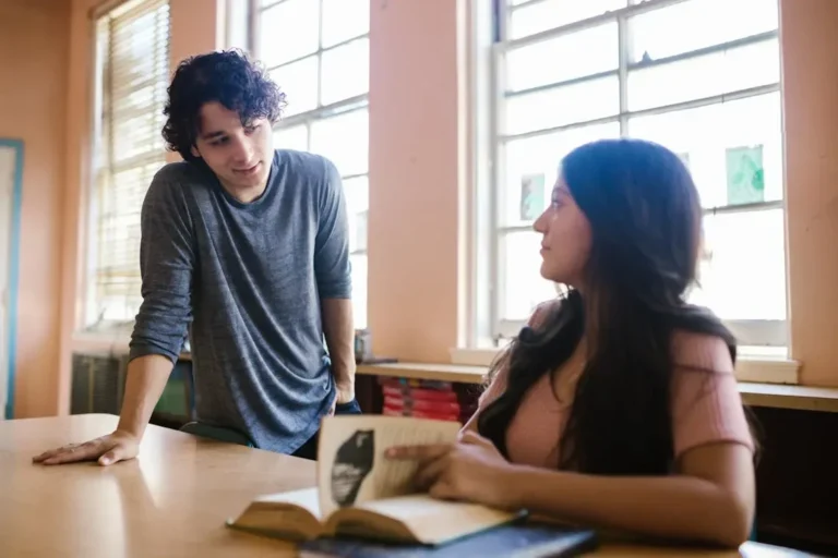 Two young adults engaged in a classroom discussion, focusing on study materials at a desk.