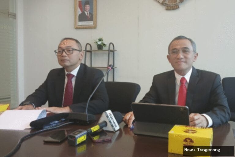 Dua pria berpakaian formal duduk di meja dengan mikrofon dan tablet.