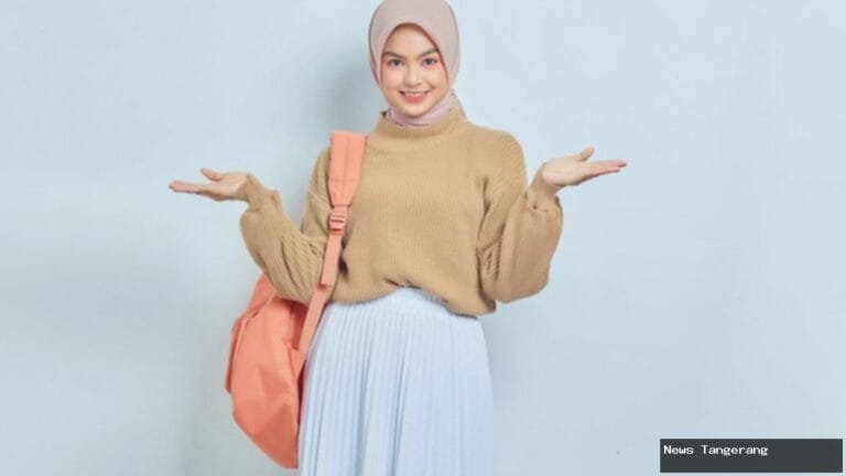 Mahasiswi berhijab dengan rok plisket, sweater, dan tas ransel tersenyum.