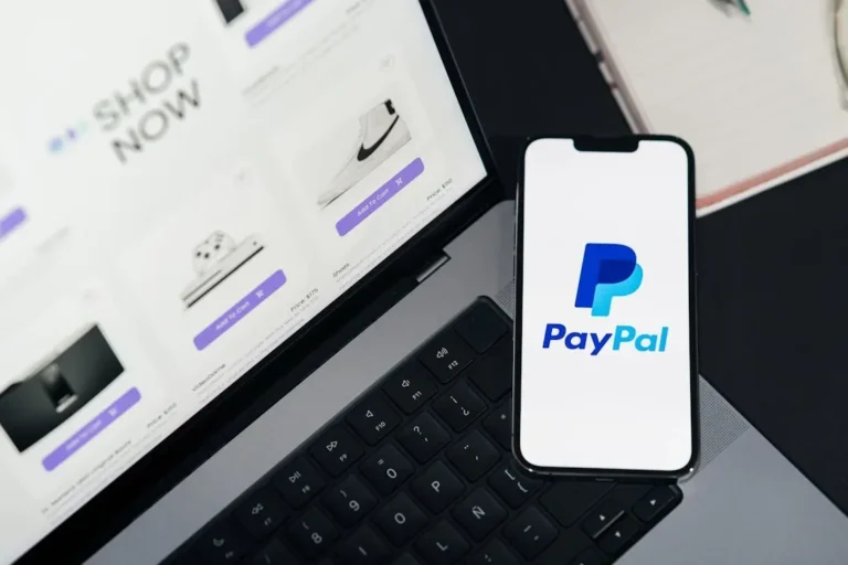Smartphone displaying PayPal logo next to laptop with online shopping site open.