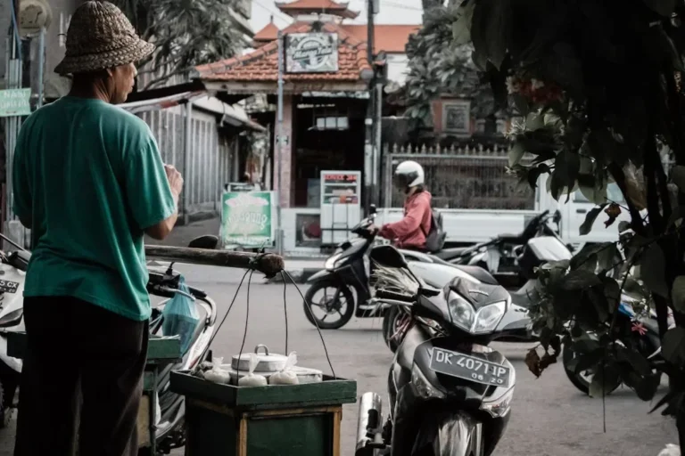 A street vendor in Marga, Bali, Indonesia, sells goods to passing motorcyclists. Busy street life scene.