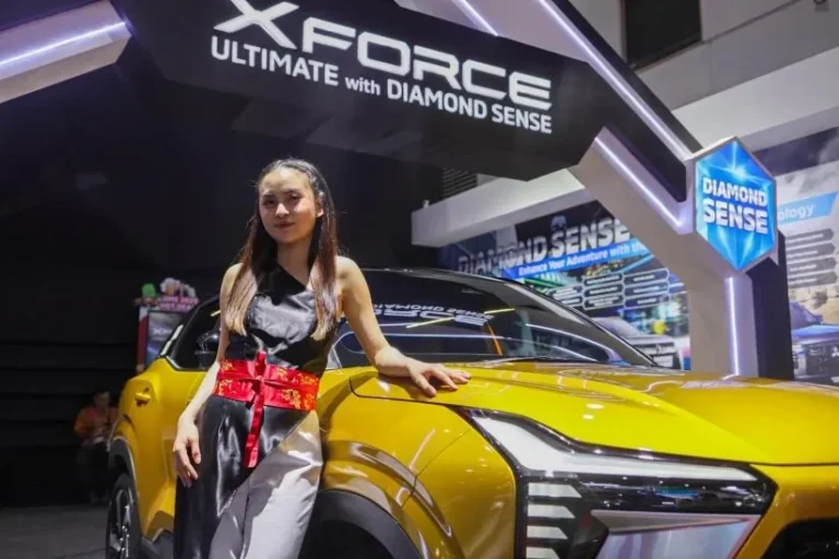 Usher IMOS 2025 berpose di samping mobil Mitsubishi XForce berwarna kuning keemasan.