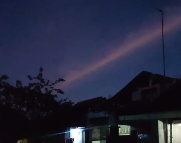 Langit malam berwarna ungu dengan awan merah muda tipis di atas atap rumah.