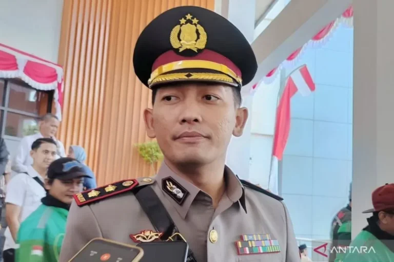 Foto close-up polisi berseragam lengkap, ekspresi wajah serius, latar belakang kerumunan.