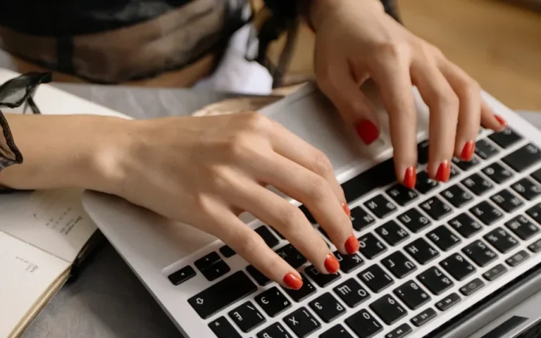 Tangan wanita mengetik di laptop, kuku merah, buku catatan di samping.