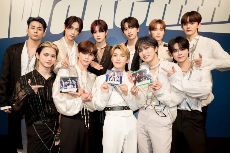 Sembilan pria muda grup musik berpose ceria memegang album "Awakening" dengan busana stylish.