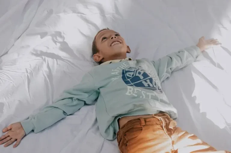 A cheerful child in a light sweater enjoys leisurely relaxation on a sunlit bed.