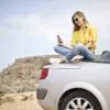 Smiling young woman sitting on a car at the beach in Mgarr, Malta using a smartphone.