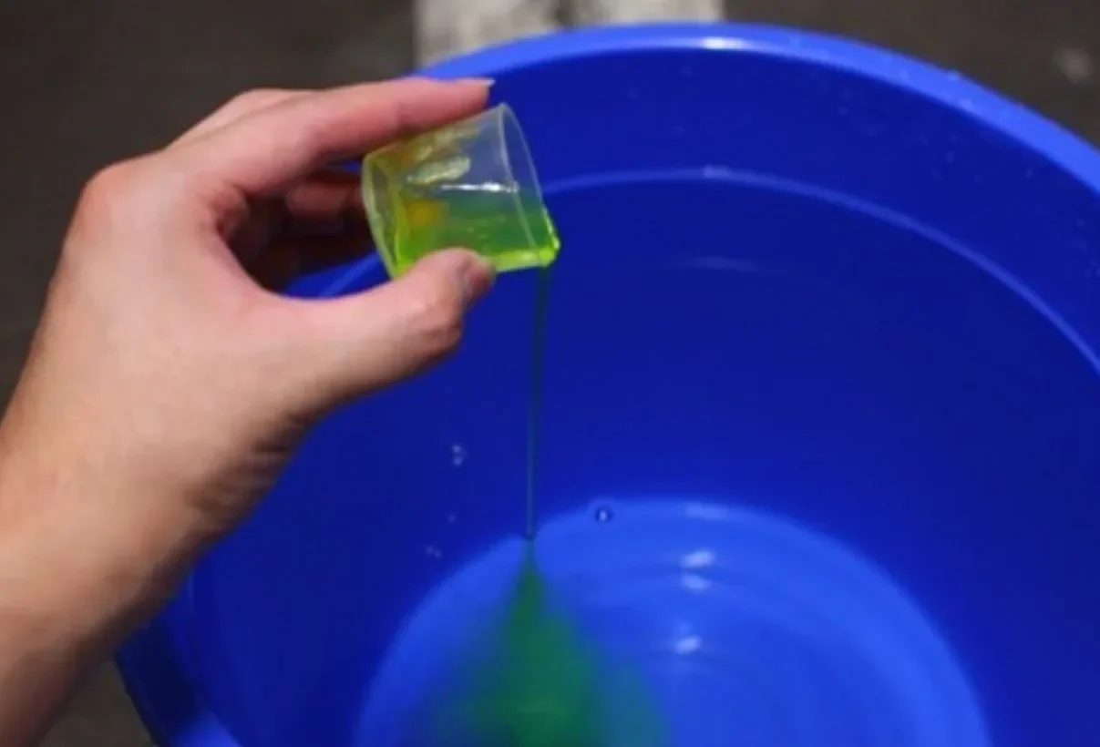 Tangan menuangkan cairan hijau dari gelas takar ke dalam ember biru.