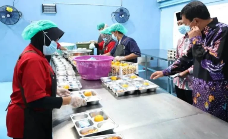 Pekerja mengemas makanan dalam baki di fasilitas dapur modern, diawasi pejabat.