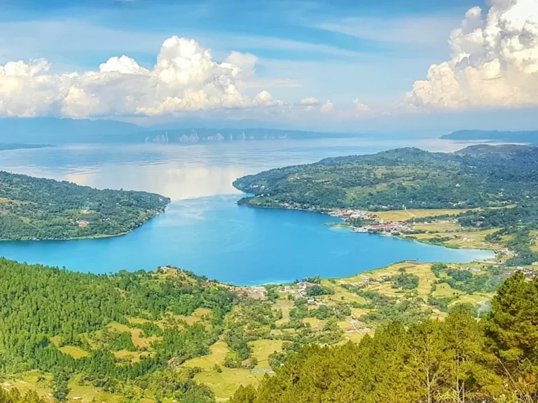 Panorama danau biru luas dikelilingi perbukitan hijau subur di bawah langit cerah berawan.