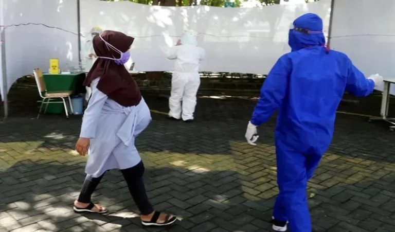 Warga mengenakan masker berjalan melewati petugas APD di area skrining atau tes kesehatan.