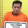 Pria tersangka berkaos oranye memegang tanda "Tes Urine Tersangka" di sebuah kantor polisi.