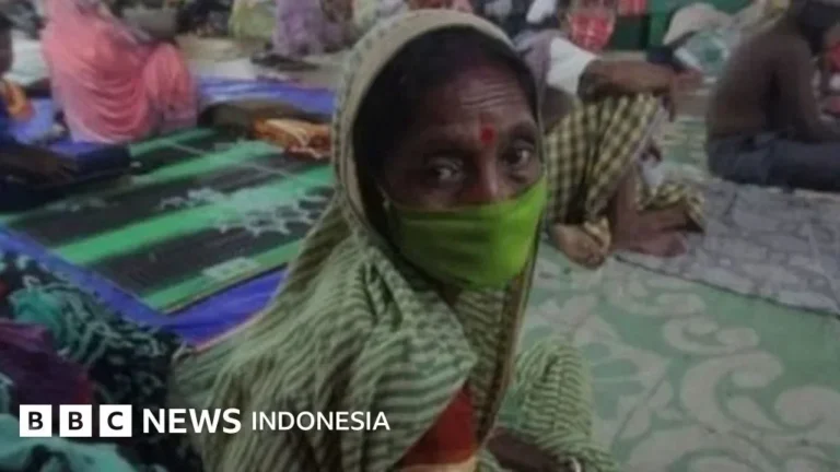 Wanita lansia berselendang dan bermasker hijau menatap kamera di tengah kerumunan orang.