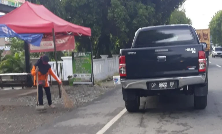 Petugas kebersihan wanita menyapu trotoar di samping jalan dengan mobil pikap melintas.