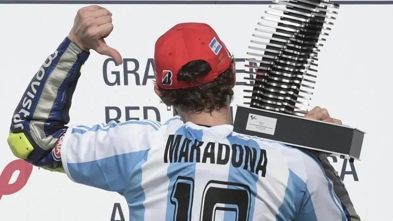 Pembalap MotoGP dengan jersey Maradona dan topi Argentina, memegang trofi sambil menunjukkan jempol ke bawah.