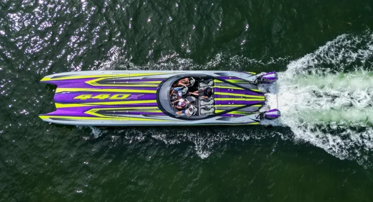 Perahu motor performa tinggi ungu-kuning dengan penumpang melaju kencang di air, dilihat dari atas.