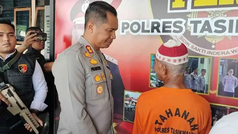 Seorang polisi menatap punggung tahanan berkaos oranye bertuliskan 'Tahanan Polres Tasikmalaya'.