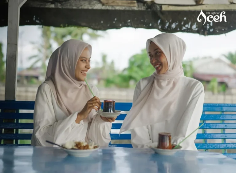 Dua wanita berhijab tersenyum bahagia sambil menikmati minuman dan makanan di kafe tepi air Aceh.