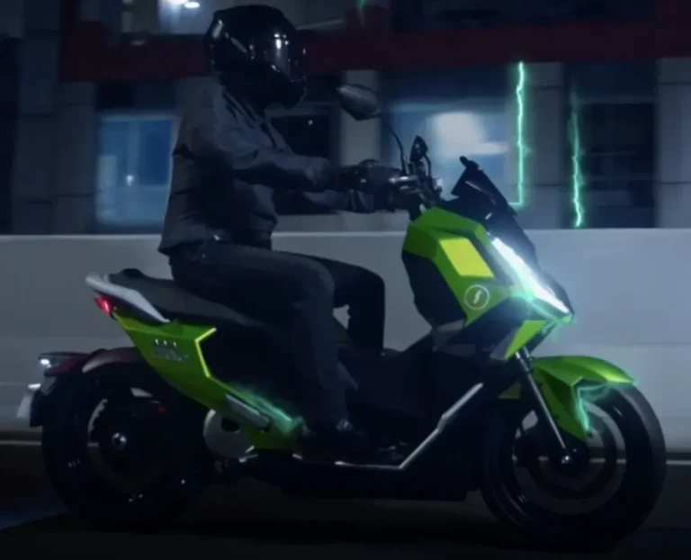 Seorang pengendara berhelm mengendarai skuter listrik hijau modern di jalan perkotaan pada malam hari.