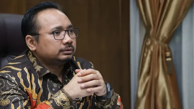 Pria berkacamata dan berbaju batik gelap duduk serius dengan tangan terkatup.