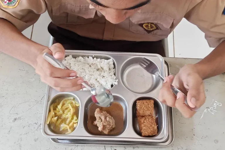 Seseorang berseragam cokelat menyantap hidangan lengkap nasi, lauk pauk, dan sayur di nampan makan.