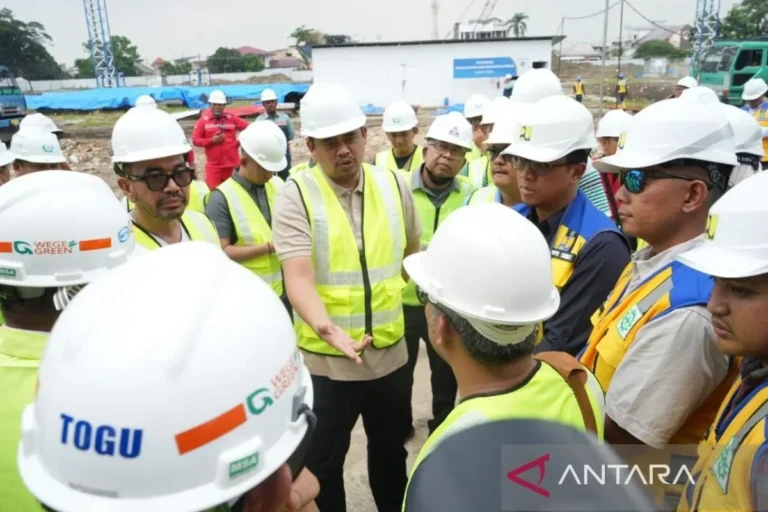 Sekelompok pejabat dan insinyur berhelm putih meninjau kemajuan proyek konstruksi di lapangan.