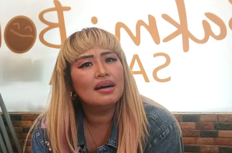 Wanita berambut pirang dengan poni, riasan mata pink, dan jaket denim tampak berbicara serius.