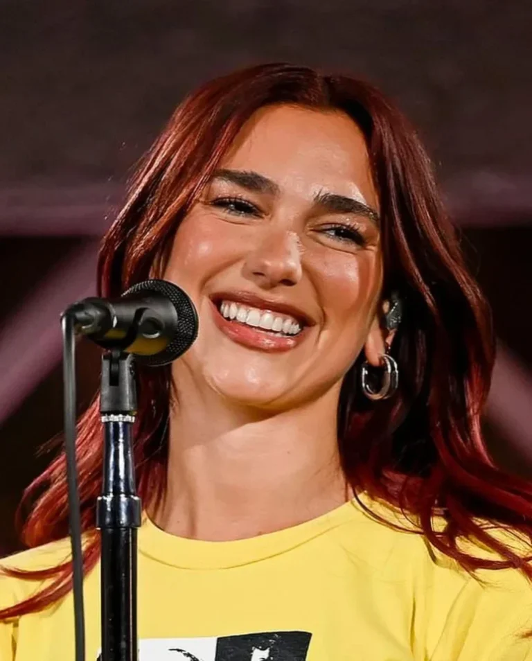 Penyanyi Dua Lipa tersenyum ceria di panggung dengan rambut merahnya di depan mikrofon.