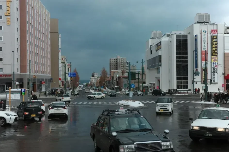 A busy winter street intersection in Otaru, Japan with cars and pedestrians on a cloudy day.